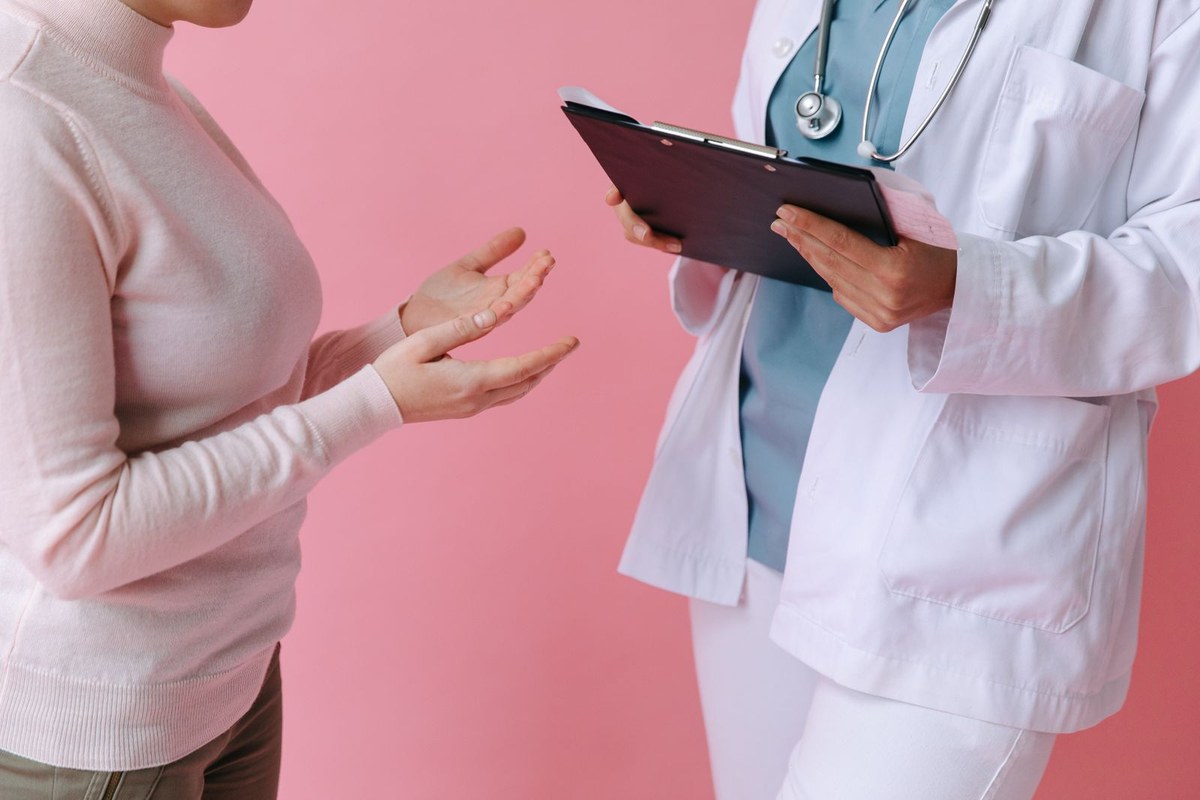 Clinician in a white coat holding a clipboard beside a patient gesturing during a consult