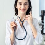 Female clinician in a white coat holding a navy stethoscope toward the camera in a bright clinic