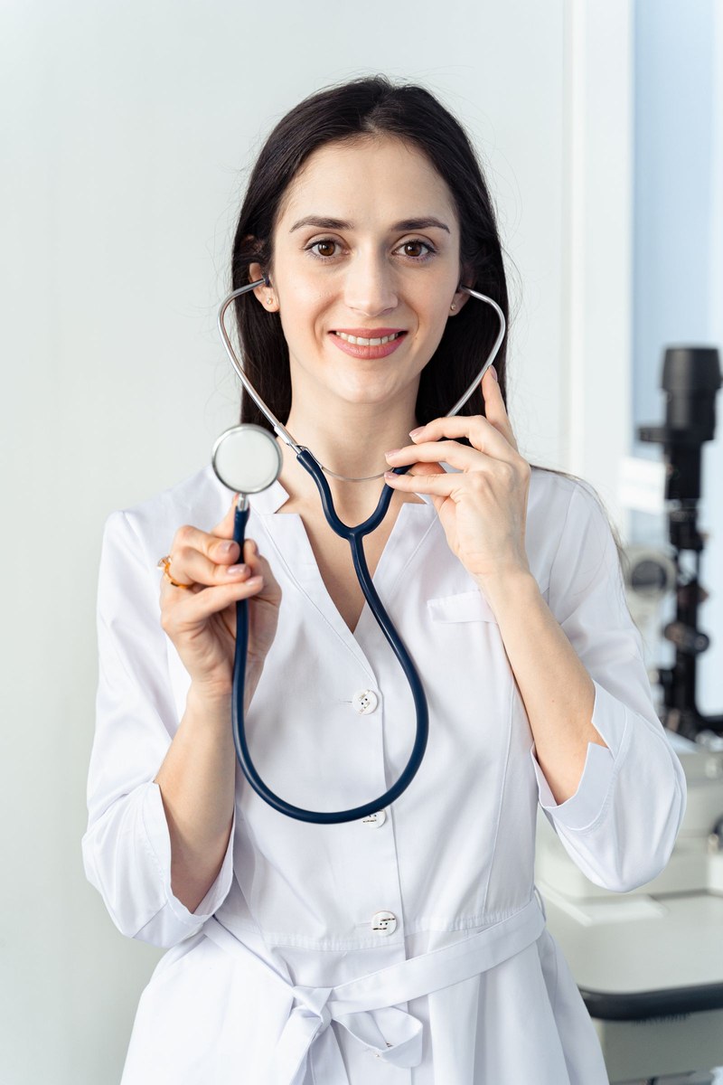 Female clinician in a white coat holding a navy stethoscope toward the camera in a bright clinic