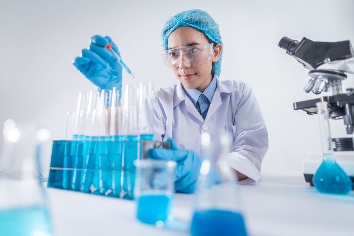 Scientist in a hair cap and gloves pipetting blue liquid into test tubes, with a microscope beside her